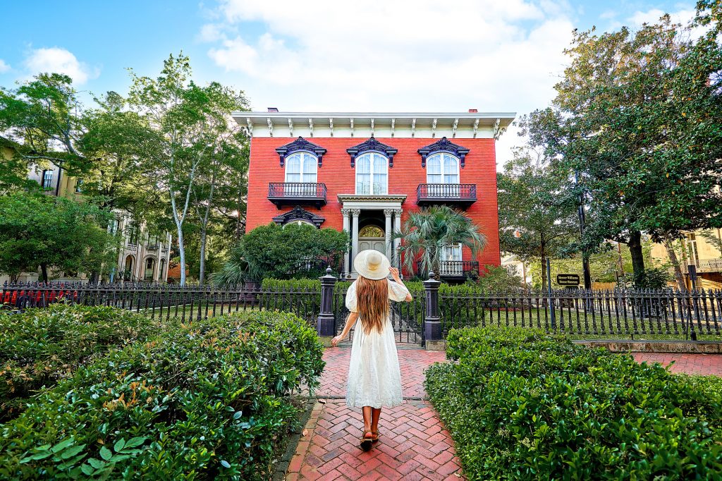 A woman in a white dress in hat standing in front of a historic brick home in a shrub garden best things to do in the usa