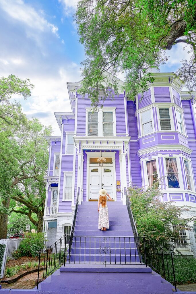 A woman in a white dress and hat standing on the steps of a historic home that is painted light purple