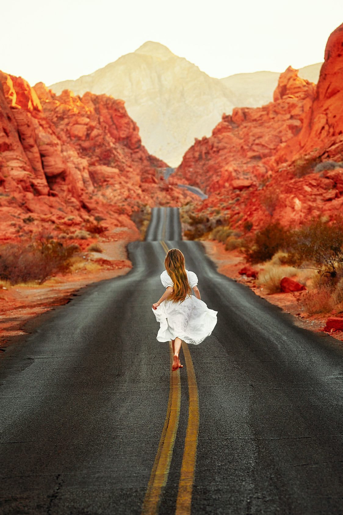 A woman in a white dress running down a road in the middle of a valley of red fire colored rock formations 