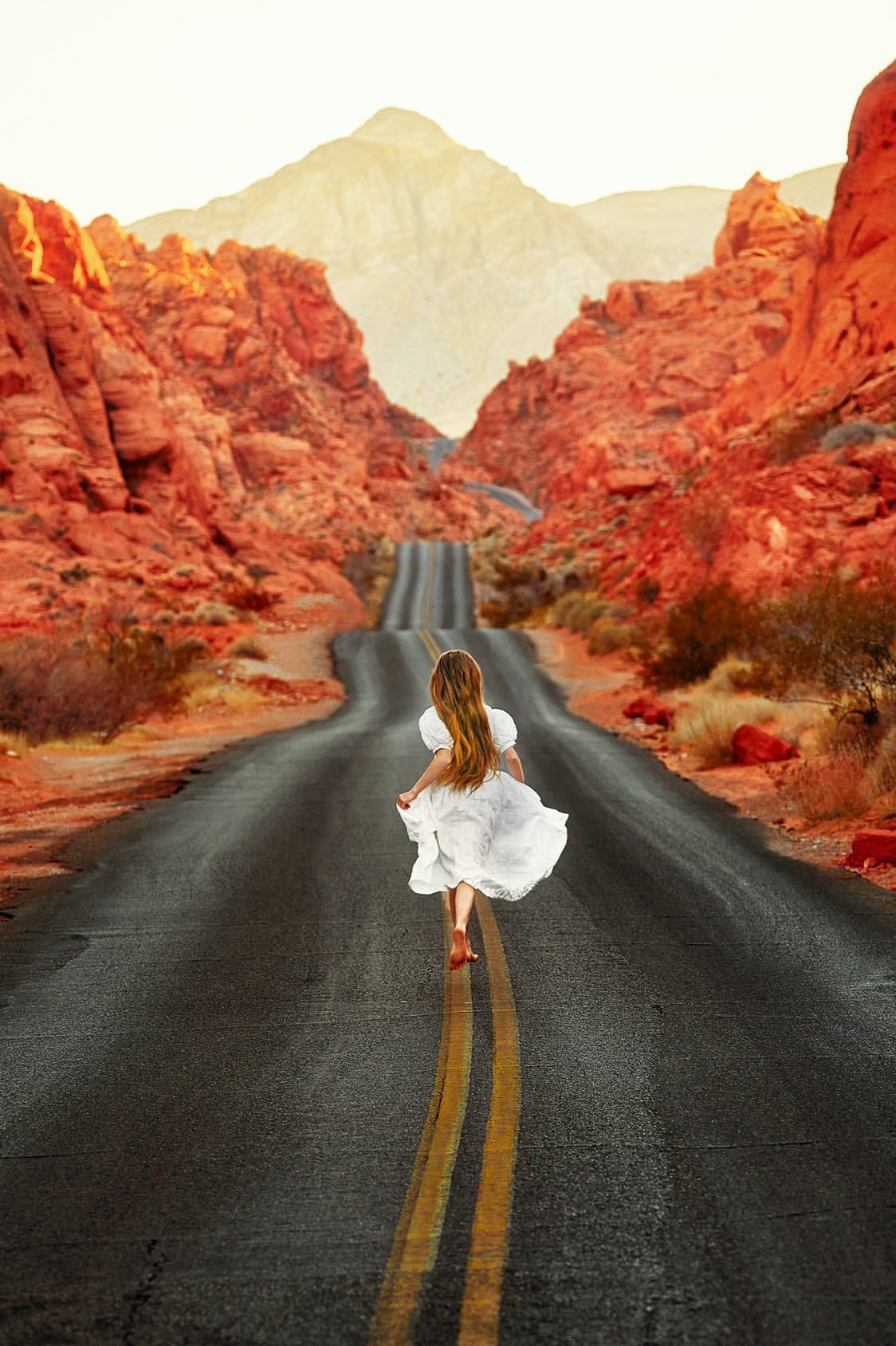 A woman in a white dress running down a road in the middle of a valley of red fire colored rock formations