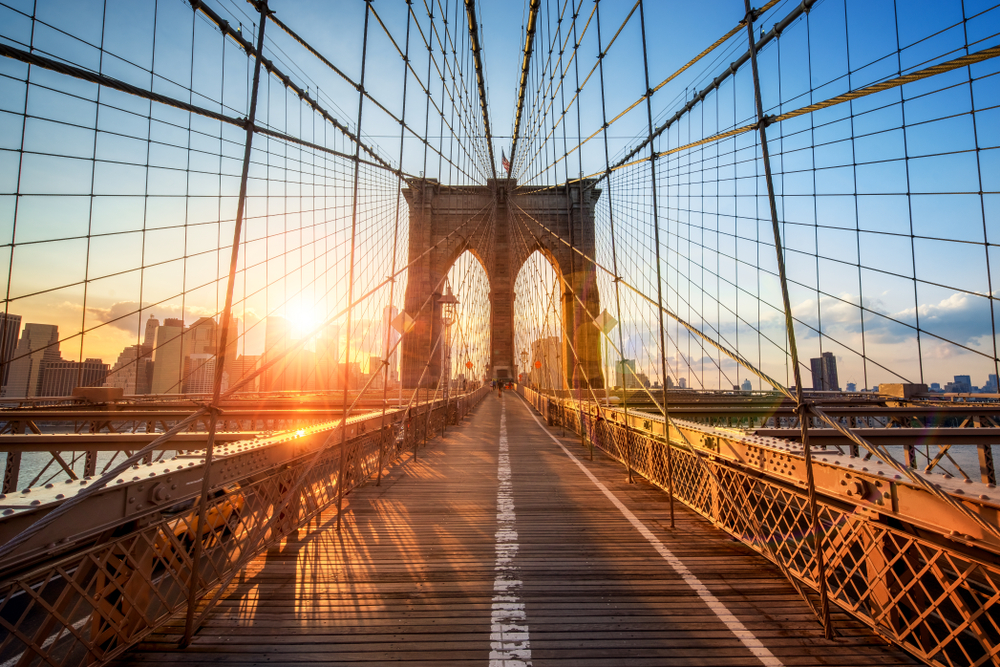 the view from on top of the Brooklyn bridge at sunset with orange glows coming in. you can see the wires on the side of the bridge and the big buildings in the back. this is a wonderful stop during your Weekend in NYC 