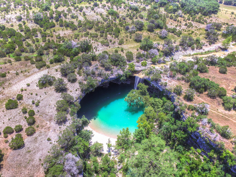 a natural swimming pool in the Dripping Springs town of Texas. the pool is big blue and clear with a waterfall cascading down one side. 