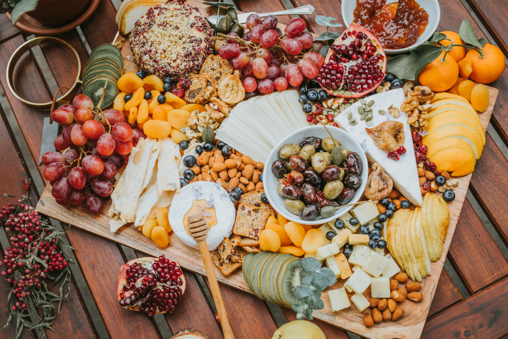 wood planked table covered in a charcuterie piece covered with rich cheeses, orange slices, apple slices, kiwi, grapes, olives and nuts galore!