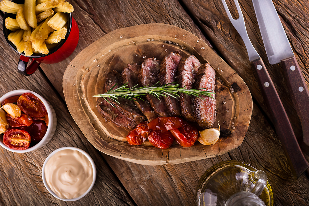 over a sliced wooden plank is beautifully cooked strips of meat with toasted tomatoes, a sprig of rosemary and chips! a traditional Brazilian barbecue!