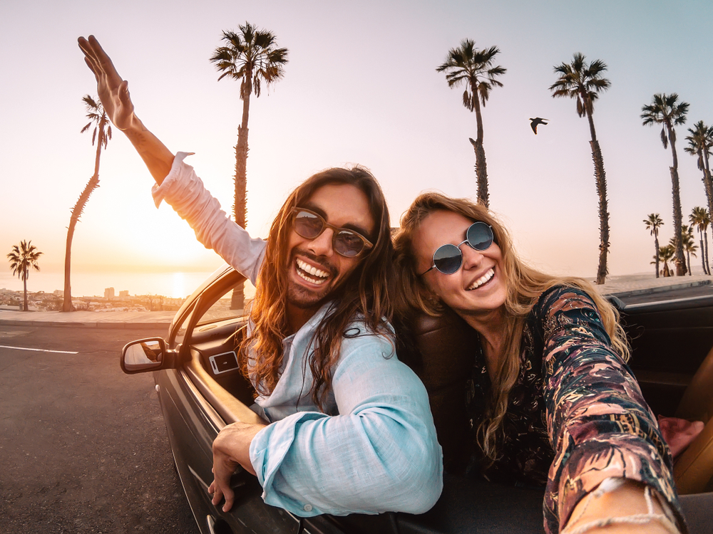 A couple in a selfie taken in a car along the beach in Florida with palm trees and the sun setting in the background