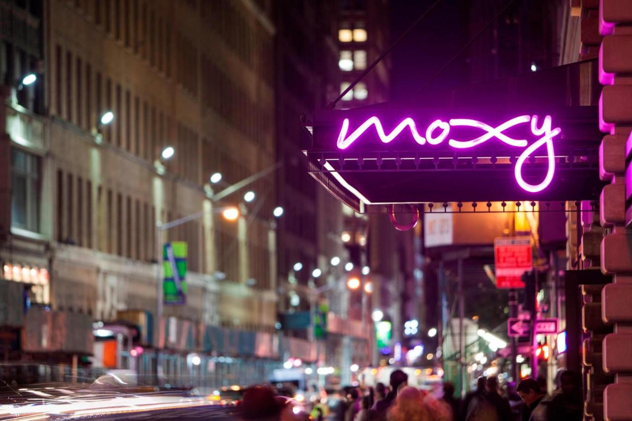the front of the Moxy Hotel in NYC Times Square the sign is lite up in a bright purple neon light 