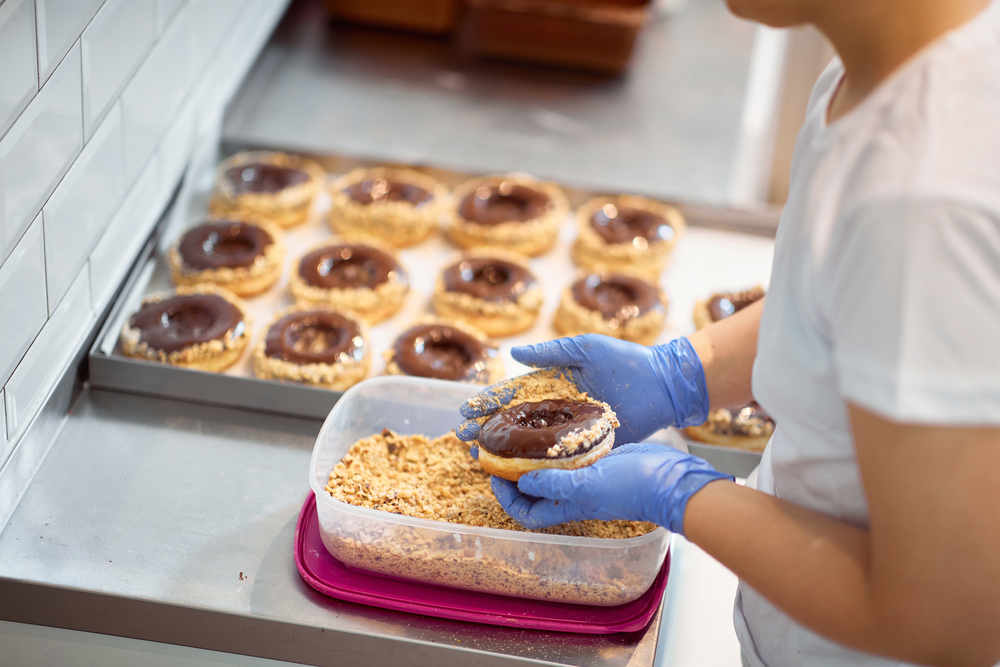 A worker with blue gloves coats a chocolate donut with something sweet and lays them on a baking pan. 