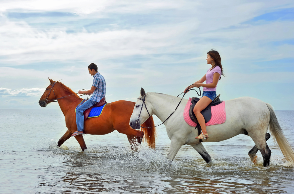 Two people ride horses through the waters on an Anna Maria beach; horseback riding through the water is offered here in Bradenton, and is one of the best things to do in Anna Maria Island.