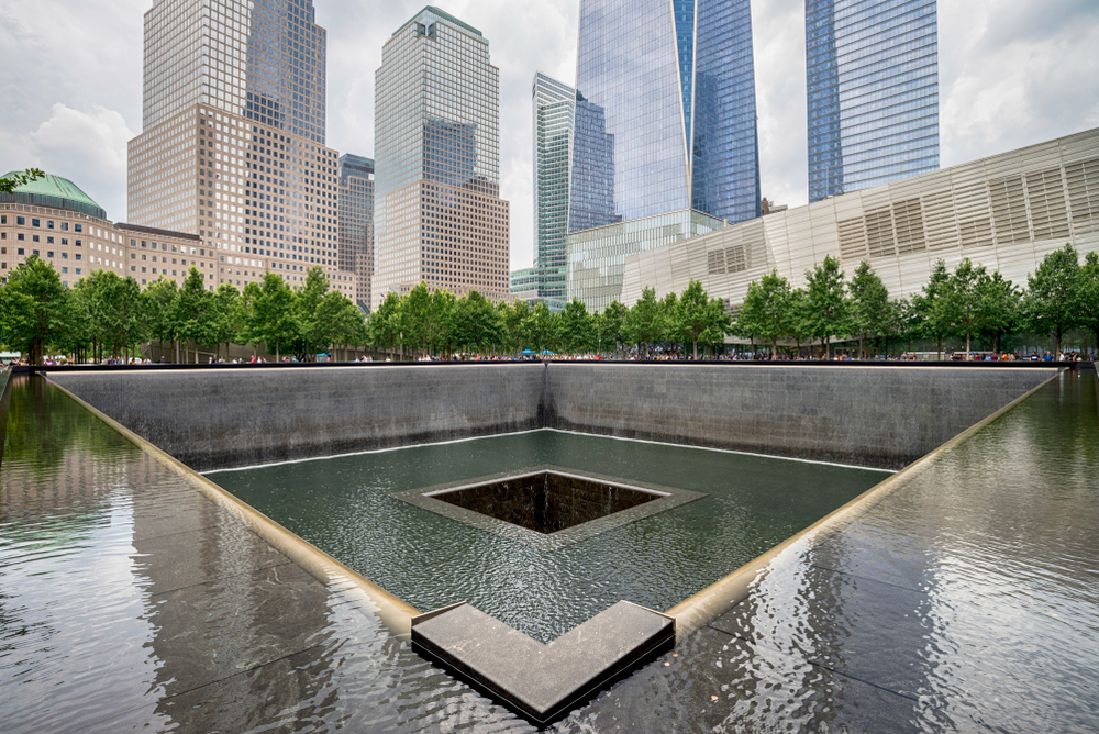 the 911 memorial in NYC with water flowing through the middle and the big buildings in the back round 