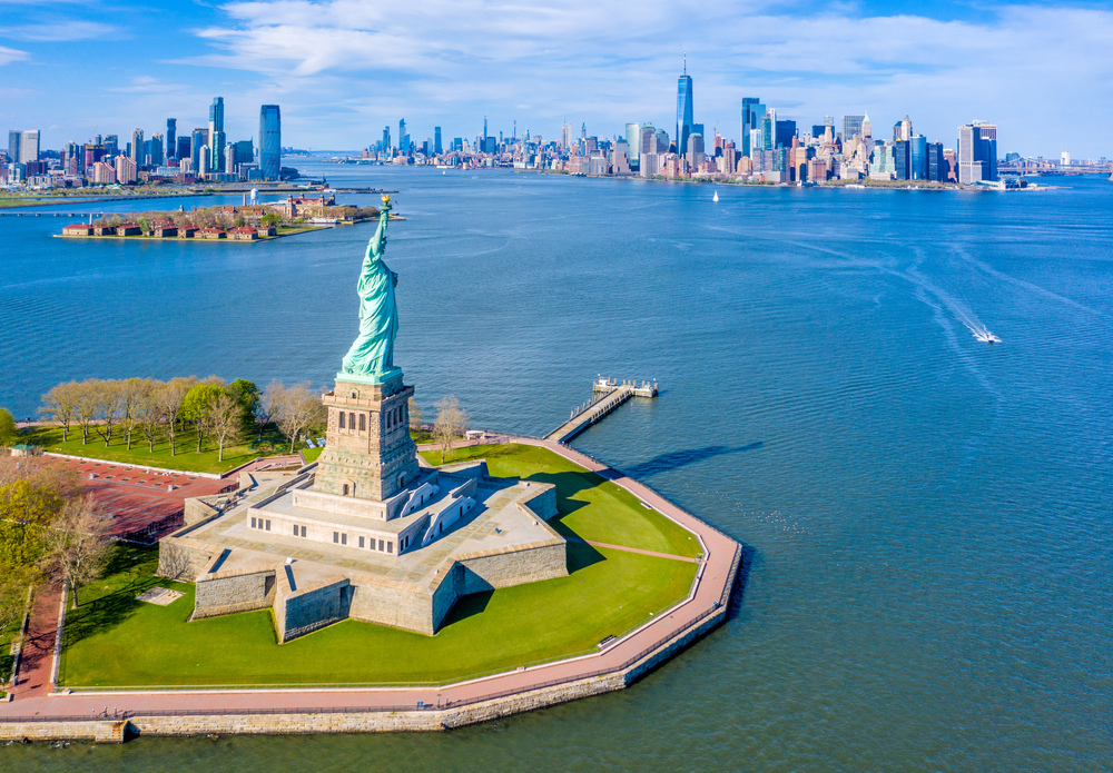 the view of a Statue of Liberty and Ellis Island from above on the Hudson River. there are a few boats going by and plenty of grass under the statue of liberty