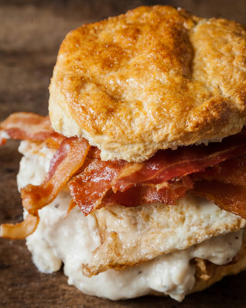 a biscuit sandwich close up photo, the sandwich has bacon, fried chicken, a fried egg, and gravy on it