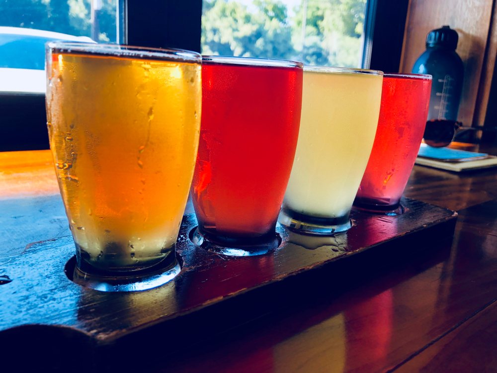 a flight of beer sits on a wooden bar in front of a window, there are four beers that are all different colors