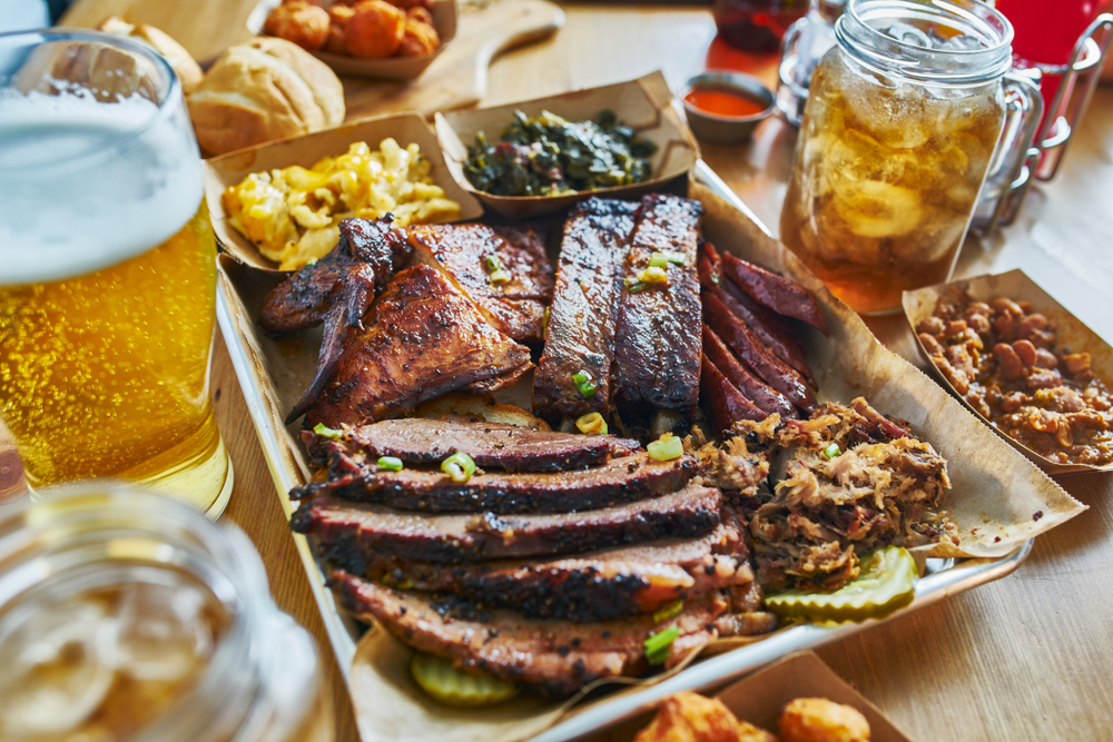 a large tray of barbecue and side dishes sits on a table next to a glass of beer and a glass of sweat tea