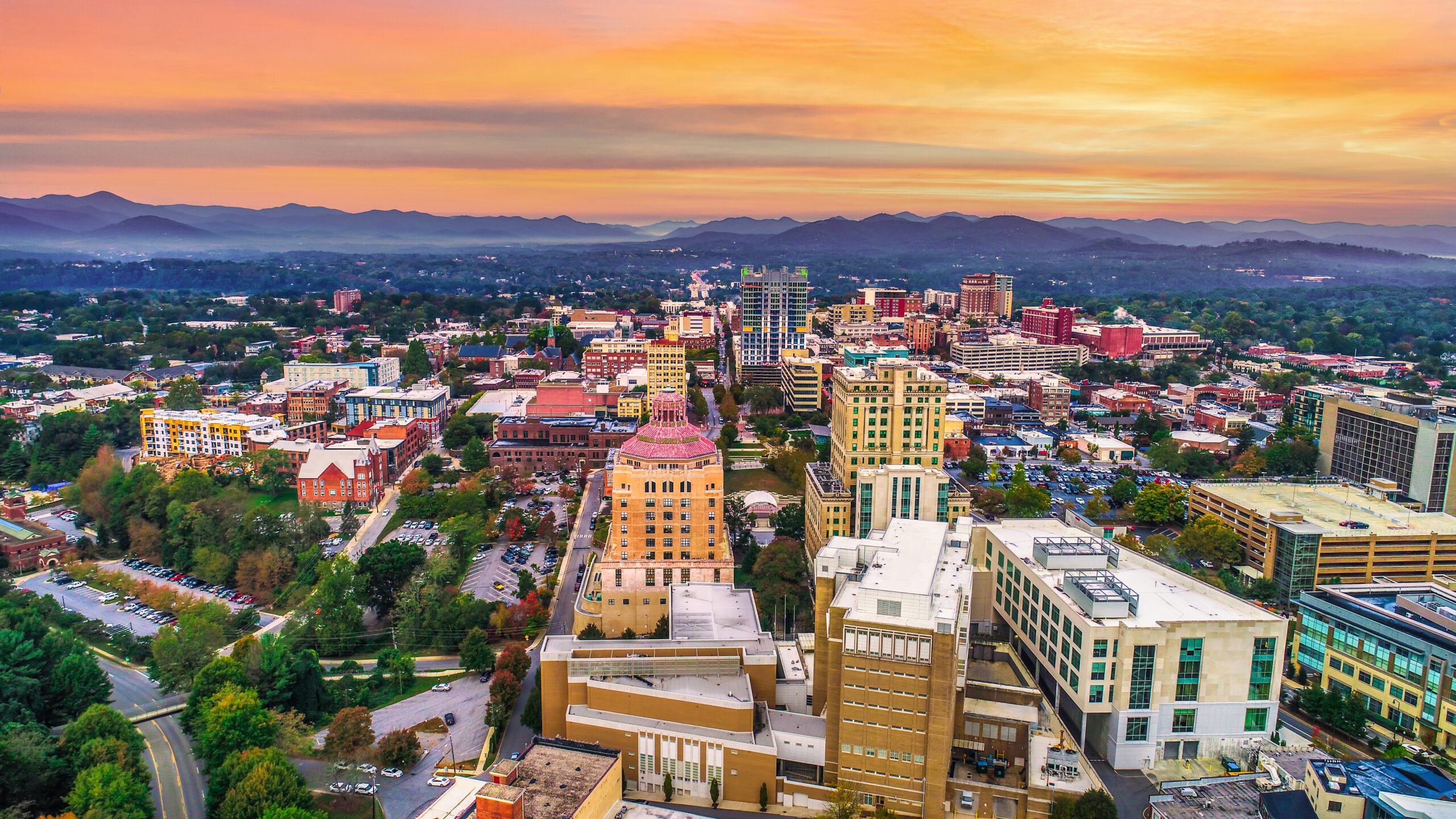 aerial photo of Asheville at sunset, there are buildings, parking lots, roads, mountains, and parks in the photo