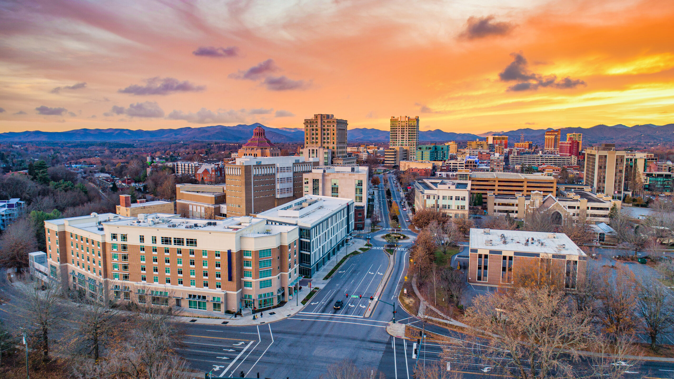 the city of Asheville at sunset, a round about can be seen in between buildings, there are mountains in the background 