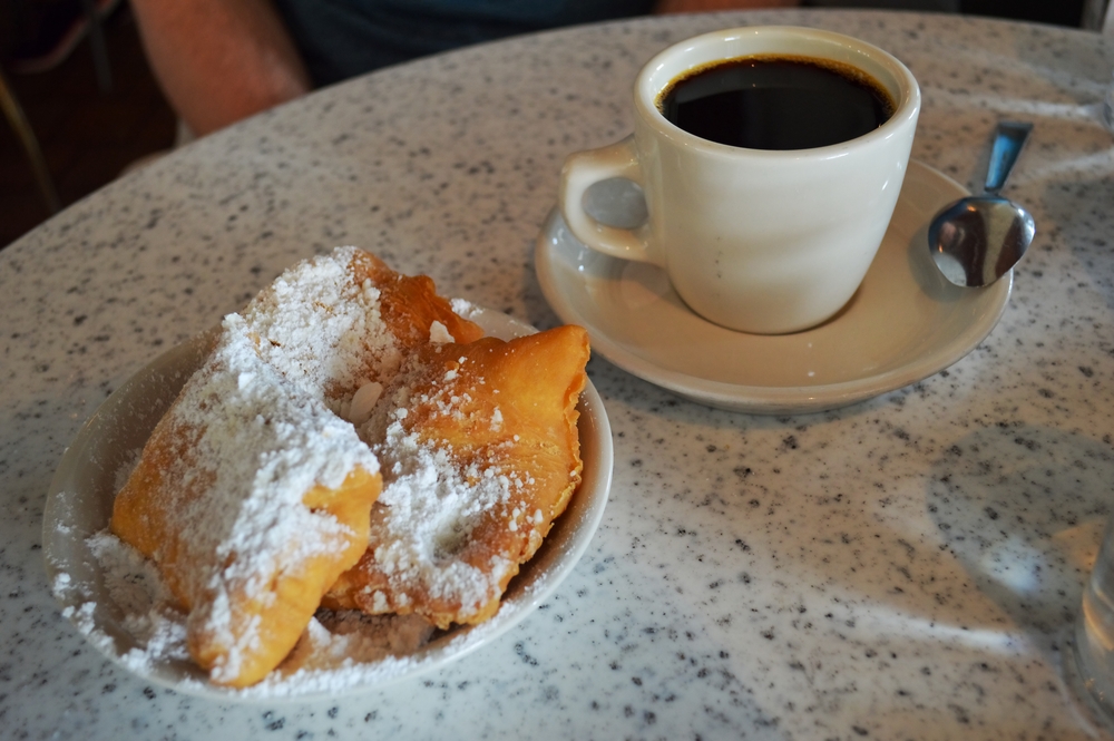 a plate of 3 beginets covered with powdered sugar and a cup of black coffee on a table