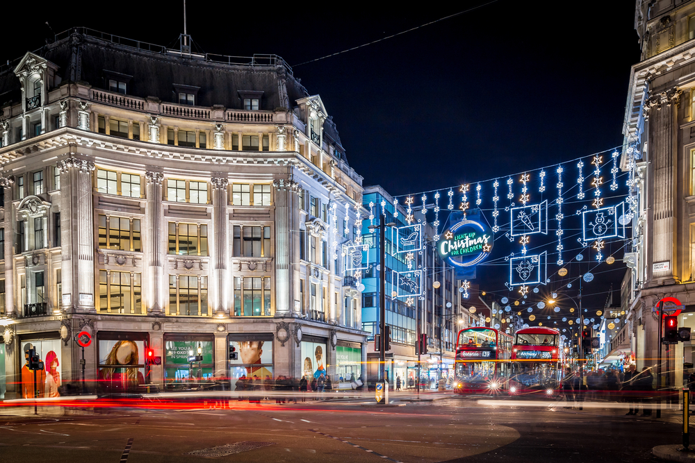 Red double decker buses driving under Christmas lights at night.