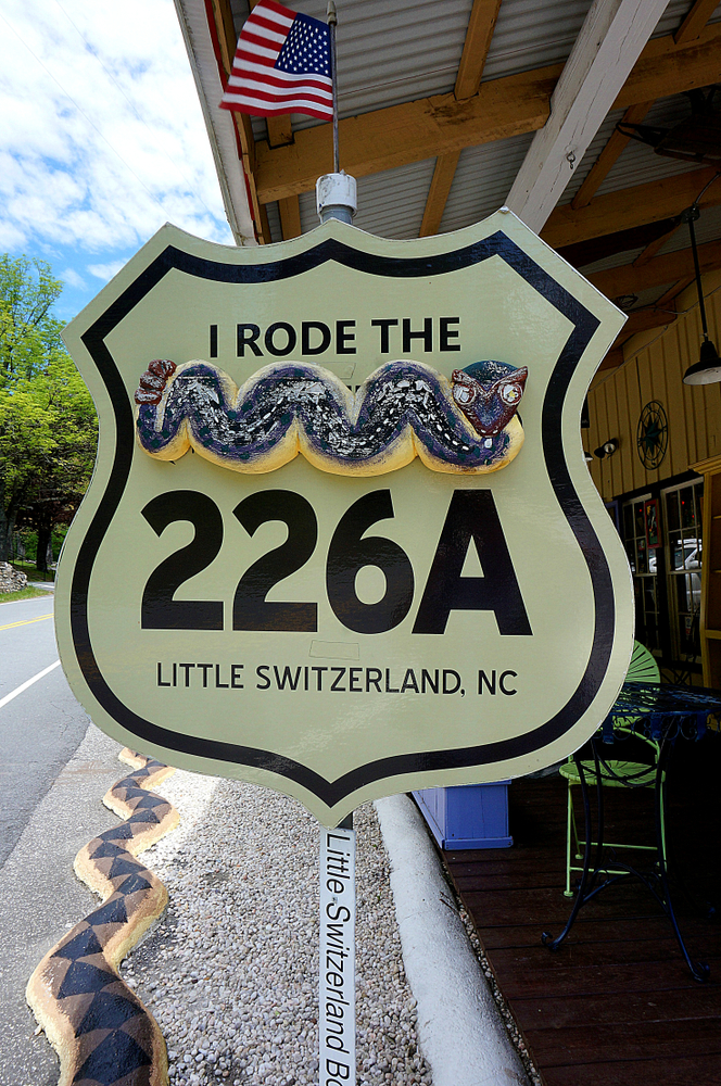 yellow highway sign marking Little Switzerland with a snake across the sign and small views of a building on the right, what you might find during one of the best weekend getaways in North Carolina 