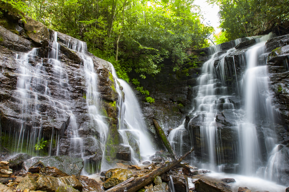 coco falls in Maggie Valley, with green trees at the top and sunlight peaking through the opening between the trees with water flowing into rocks and sticks 