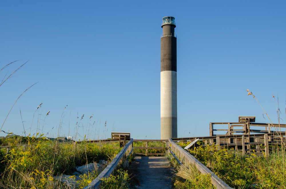 pier leading up to the oak island light house in Brunswick county with green plants growing around the walkway and a clear blue sky 
