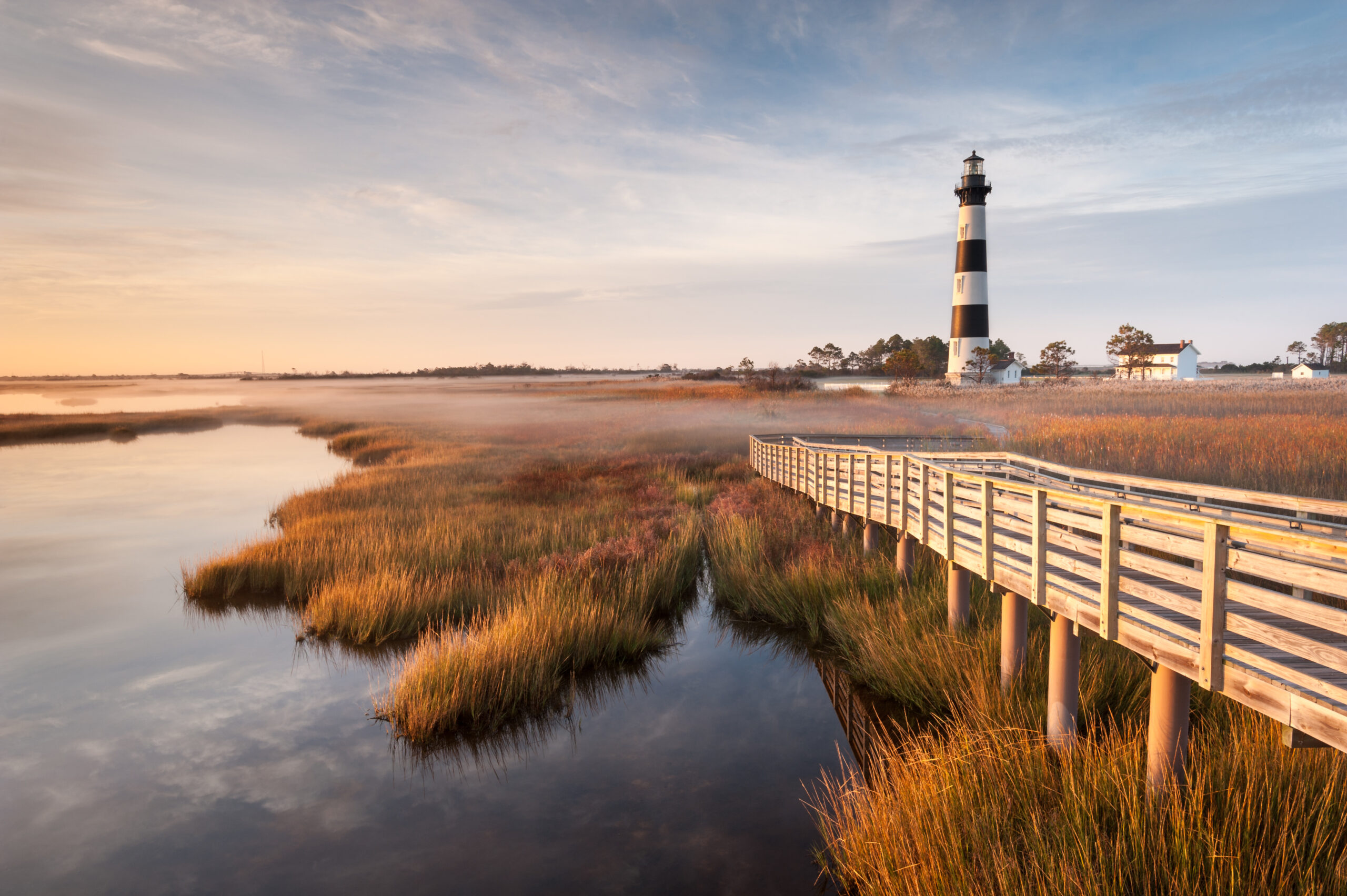 a pier next to a lighthouse on the right side near a White House and the water on the left at dusk, what you will find in the outer banks during one of the best weekend getaways in North Carolina 