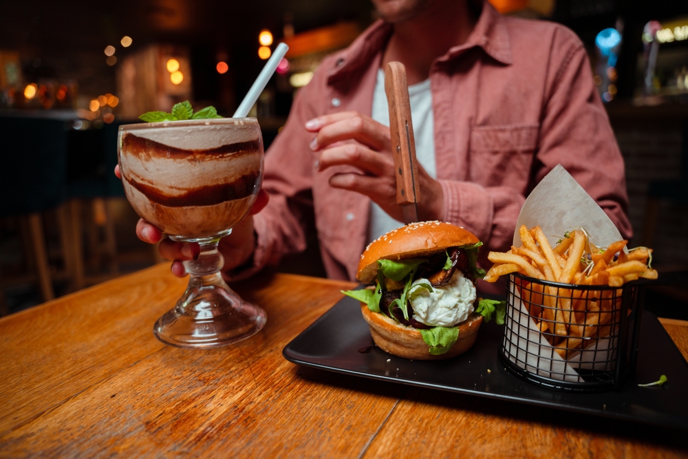 A person reaching for a large chocolate mint milkshake with a huge burger and a basket of french fries on the table