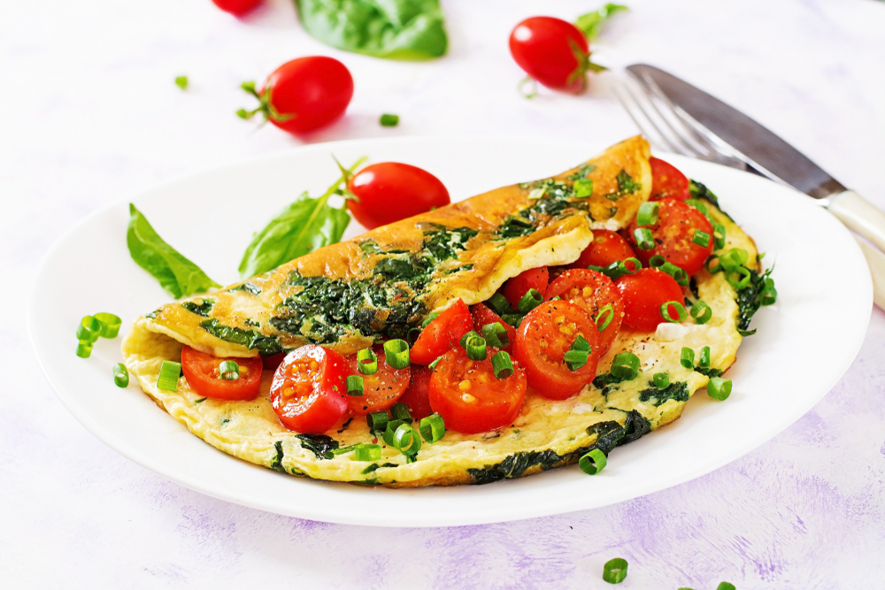 An omlette on a white plate stuffed with cheese, spinach, and tomatoes