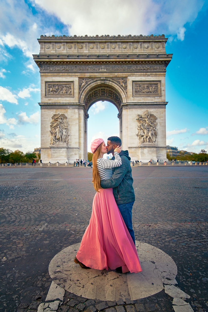 a couple kissing infront of the Arc de triomphe looking down the Champs Elysees