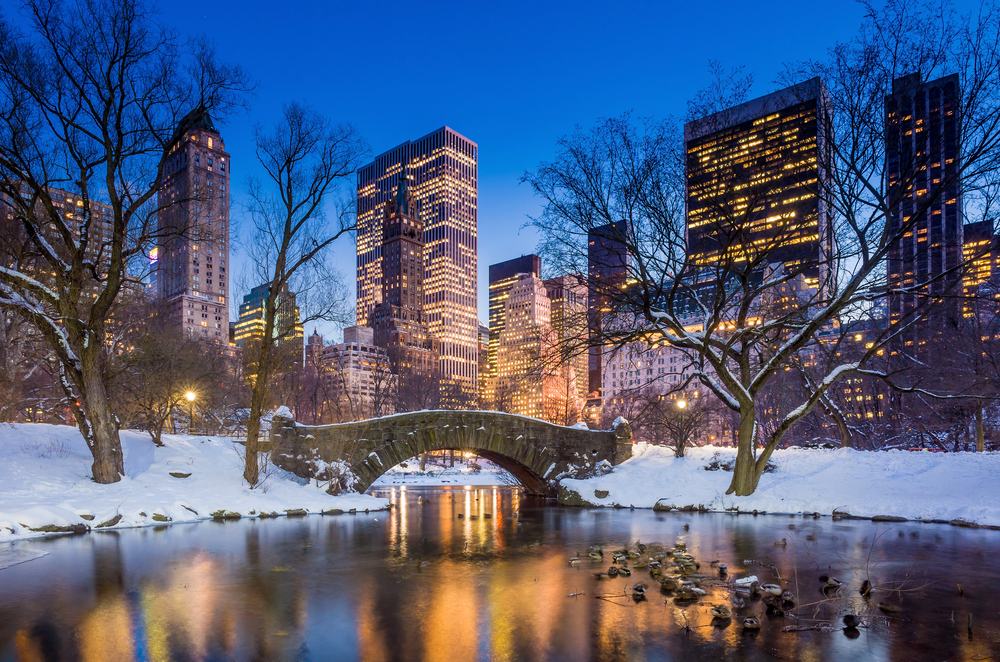 Dusk over a stone bridge in a snowy Central Park with the lit up NYC skyline in the background.