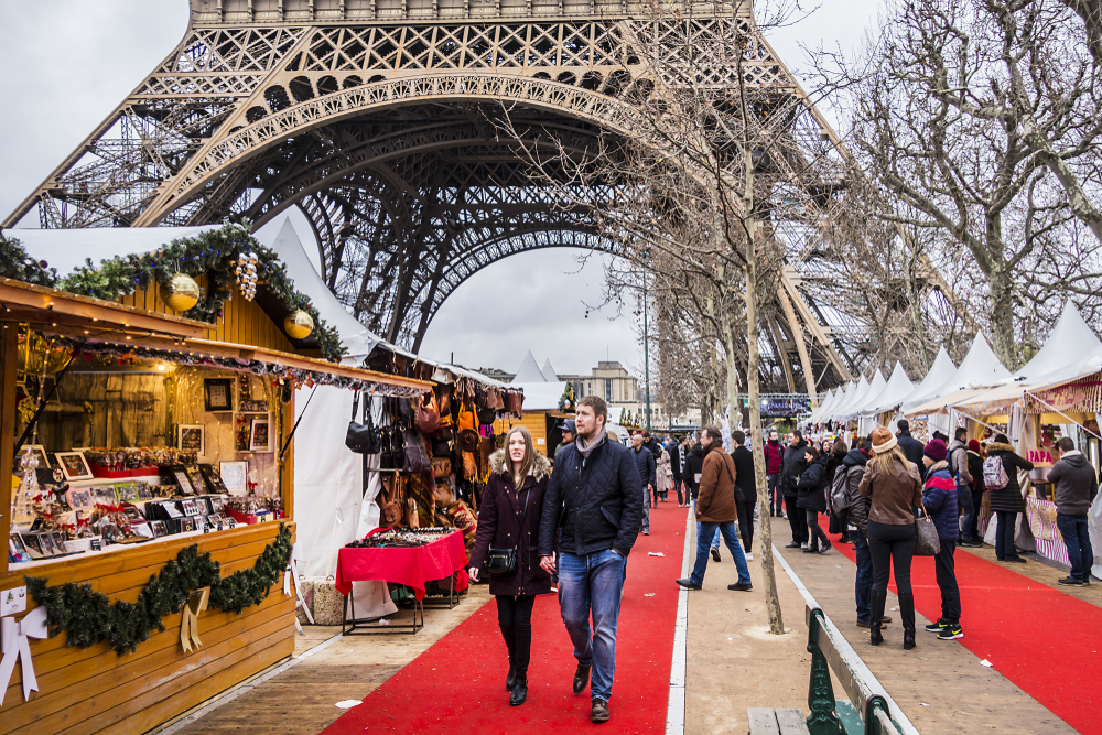 People walking around Christmas market stalls under the Eiffel Tower in Paris in winter.