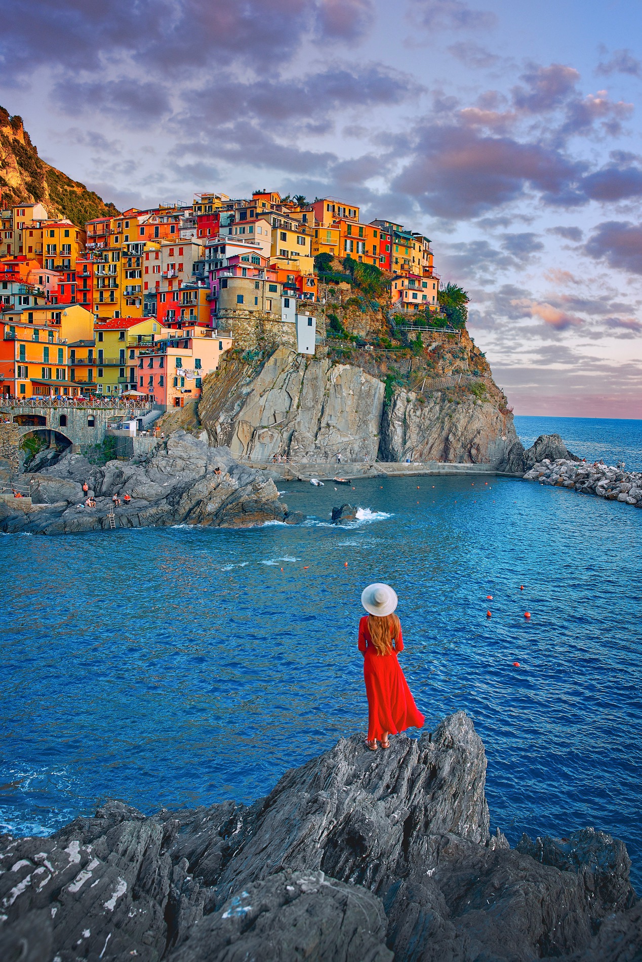 Woman in red dress and sun hat stands on a rugged rock looking across the water to a colorful village in Cinque Terre.