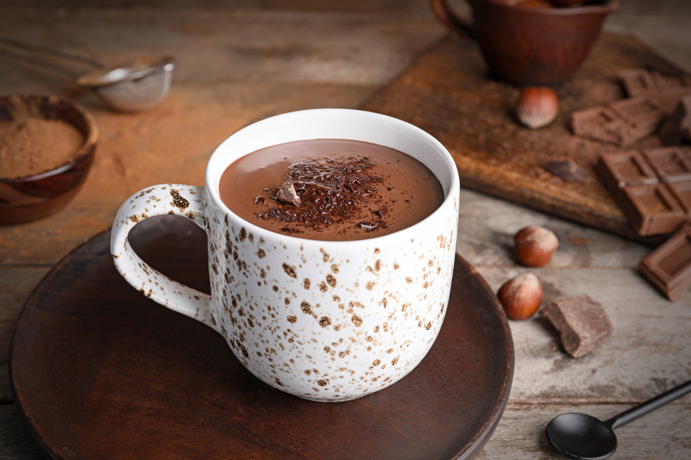 A white and brown mug with hot chocolate on a wooden table surrounded by ingredients.