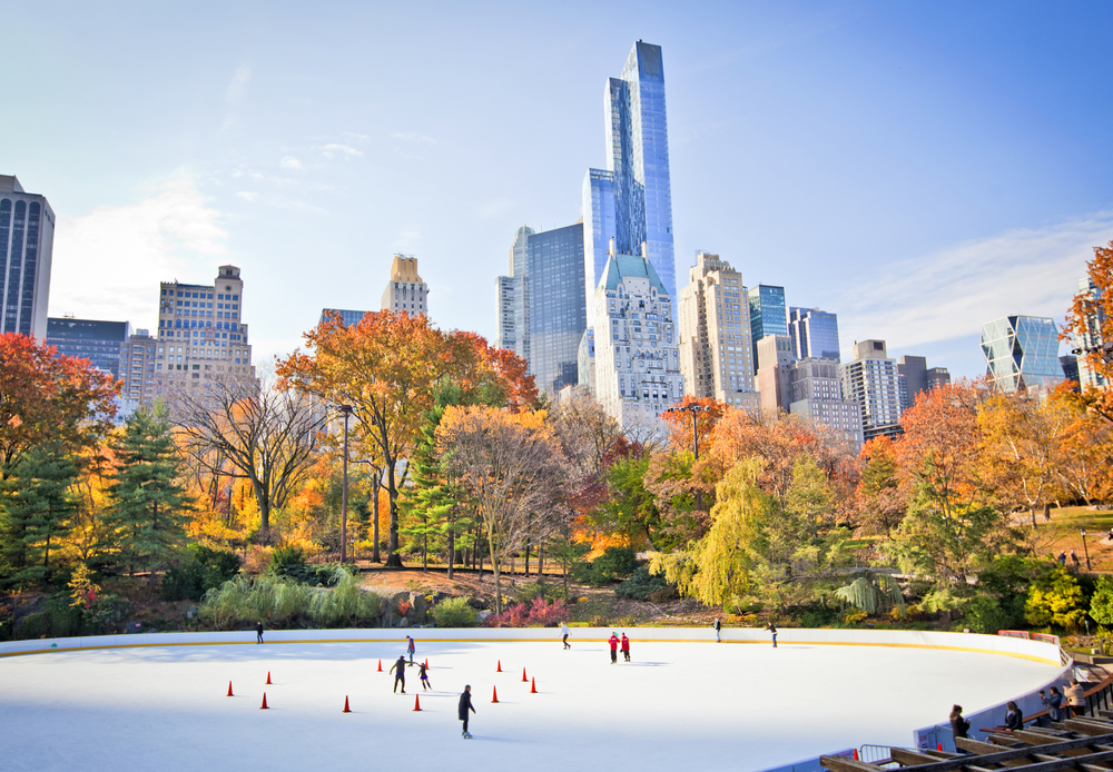 Ice skaters on a sunny day on an ice rink in Central Park next to fall trees with the skyline in the background.