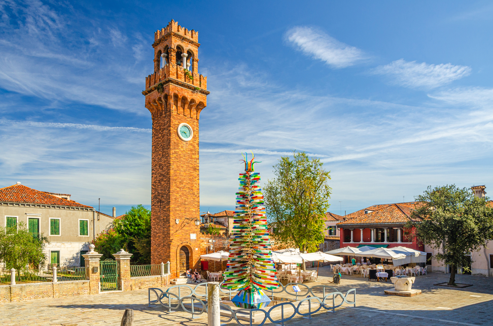 A glass Christmas tree in a square with a brick clock tower in Murano.
