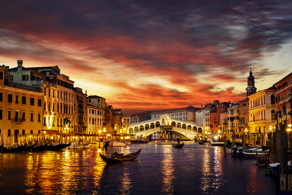 A vivid sunset over the lit up Rialto Bridge with gondolas on the canal.