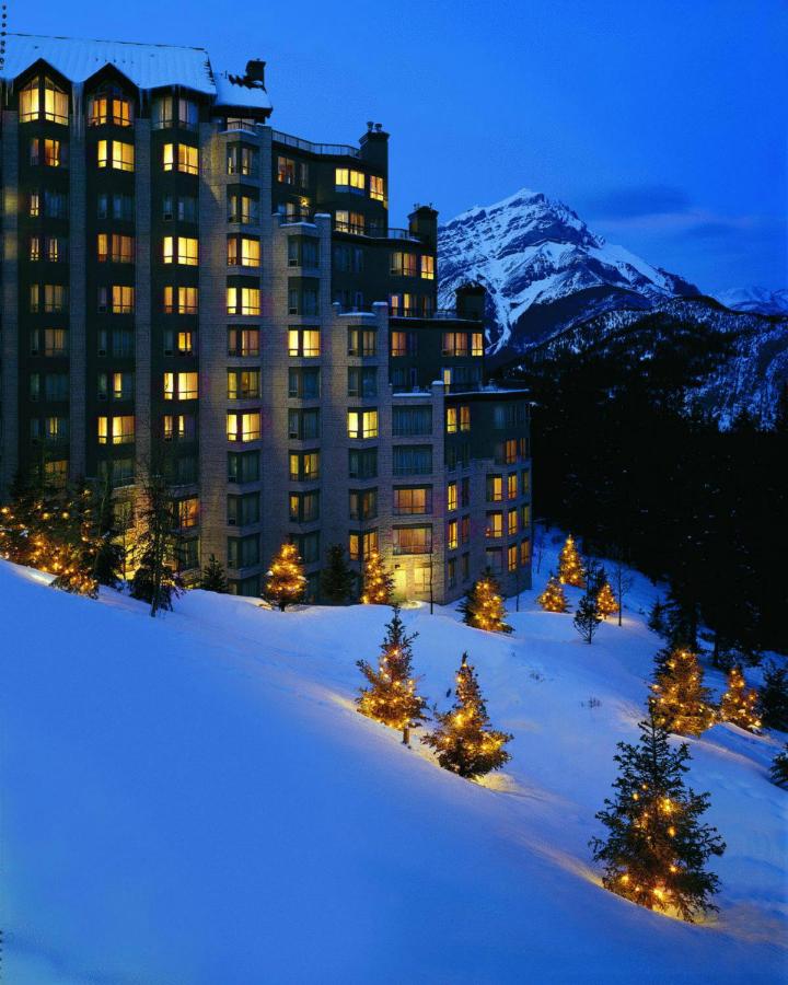 The tall Rimrock Resort Hotel at dusk with light pine trees on a snowy hill and mountains in the distance.