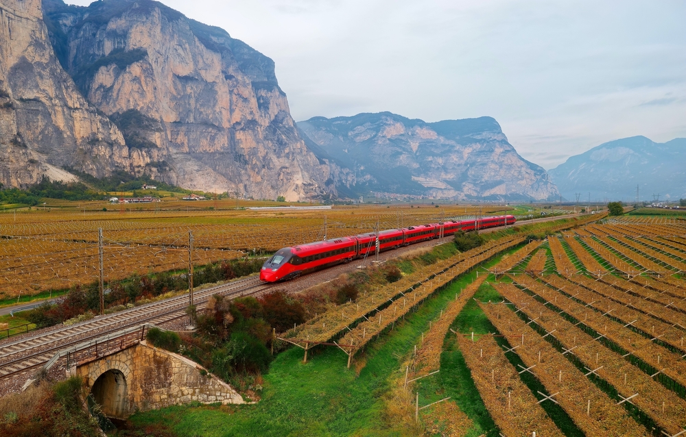 Aerial image of a red train cutting through farmland near mountains in Italy.