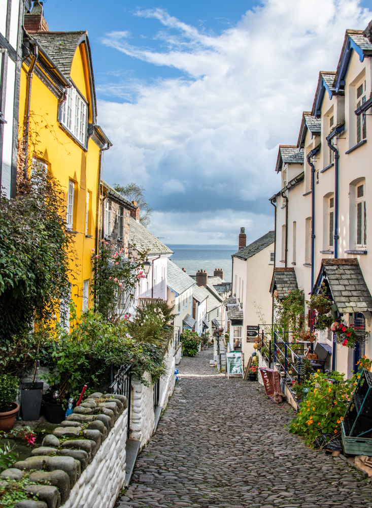 View down a street to the sea. There is a yellow house in the foreground and the other houses are white. There are lots of flowers outside.  
