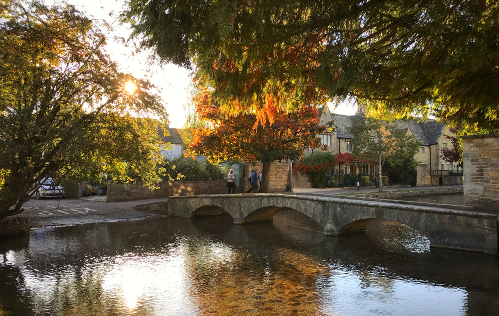 Bourton-on-the-Water one of the english villages in the autumn during the golden hour. You can see a bridge and old cottages with trees in the foreground. 