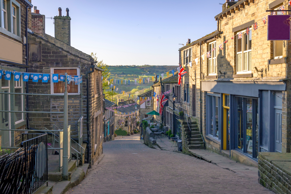 A view down Haworth town, their is coutryside in the background. The houses are made of yorkshire stone and there is bunting up between the shops.   