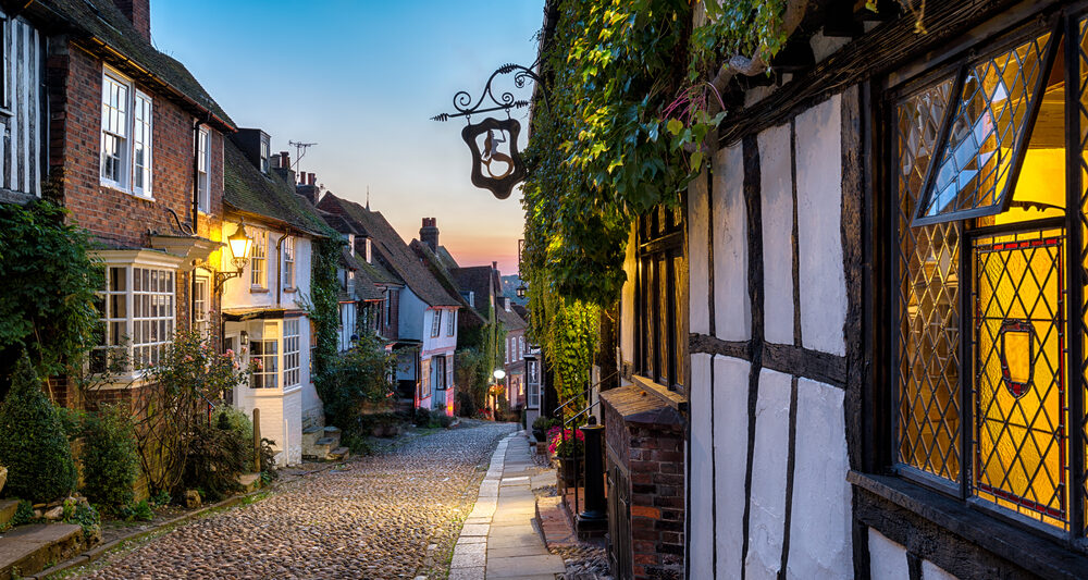 A charming cobblestone street in an old town of Rye England at dusk, flanked by traditional timber-framed buildings with warm glowing windows, inviting a sense of historical enchantment.