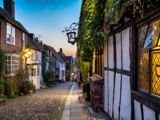 A charming cobblestone street in an old town of Rye England at dusk, flanked by traditional timber-framed buildings with warm glowing windows, inviting a sense of historical enchantment.