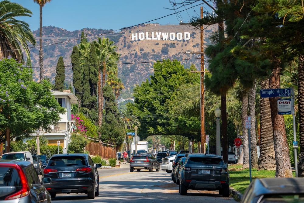 Cars moving on street amidst beautiful lush trees leading towards Mount Lee with Hollywood sign with sky in background during sunny day