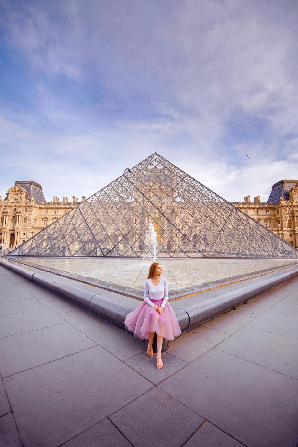 A girl in a pink skirt sitting in front of the glass pyramids in front of the Louvre one of the museums to see on your 1 day in Paris itinerary 