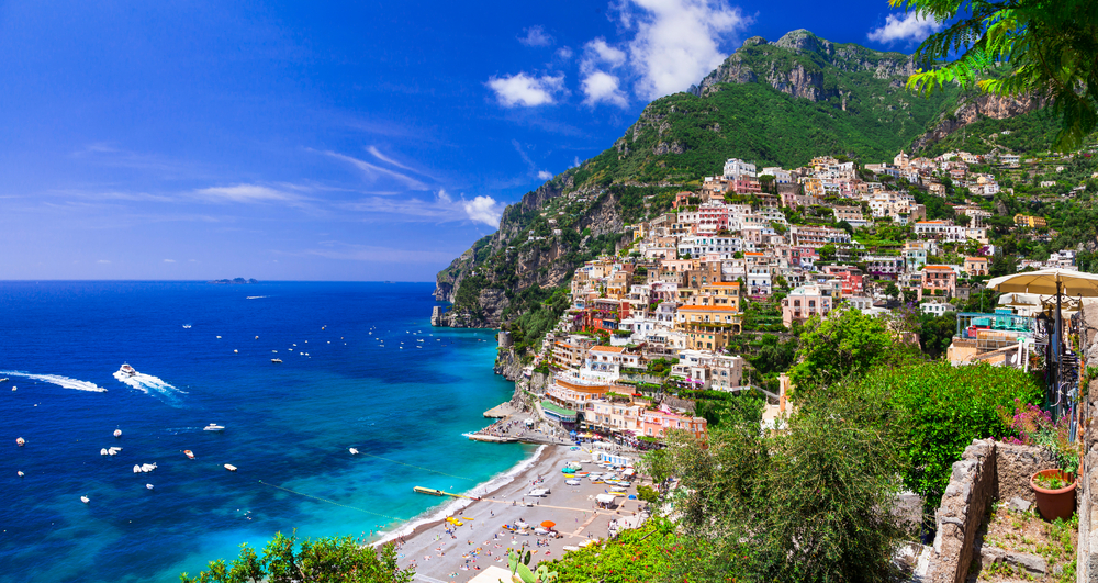 a photo of the Amalfi Coast from a balcony. there are hundreds of houses built on the hill and a small beach front with boats and beach chairs in the water. 