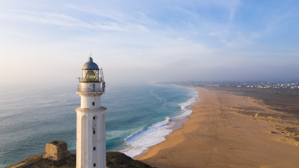 a view of a beach town in Spain with nobody on the beach and an old white light house in the front of the frame. in the distance you can see a seaside town. 