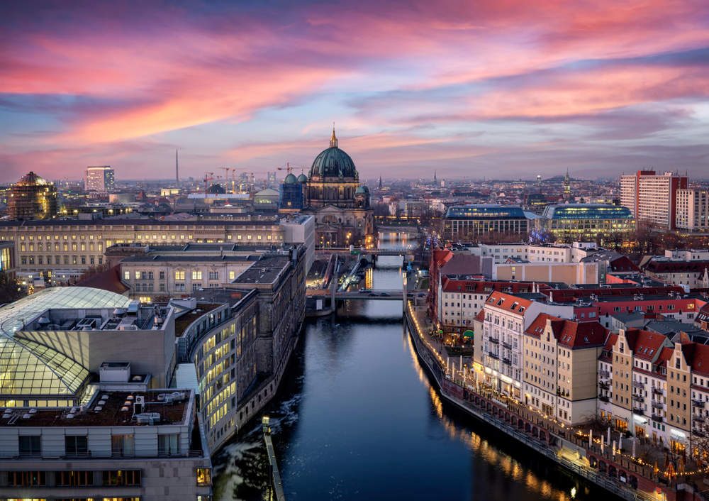 a lovely view of the old town of Berlin Germany during sunrise. the sky is a mix of purples, blues, and yellow. the cities lights are turning on and the canal is quiet and calm. 