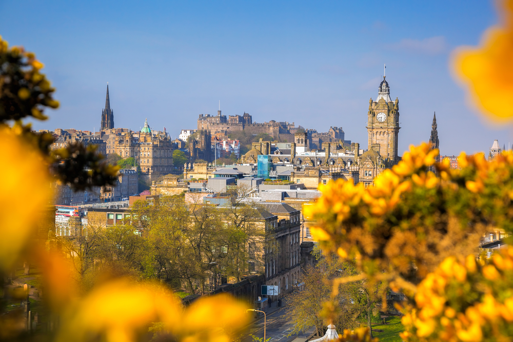 a photo of Edinburgh Scotland from the distance with big yellow flowers and the sky is perfectly blue. you can see plenty of church towers and a castle in the back round. this is one of the best places to visit in Europe in April 