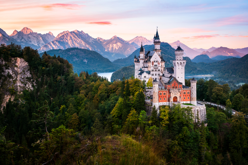 Sunset at over the Neuschwanstein Castle surrounded by trees and mountains in the distance.
