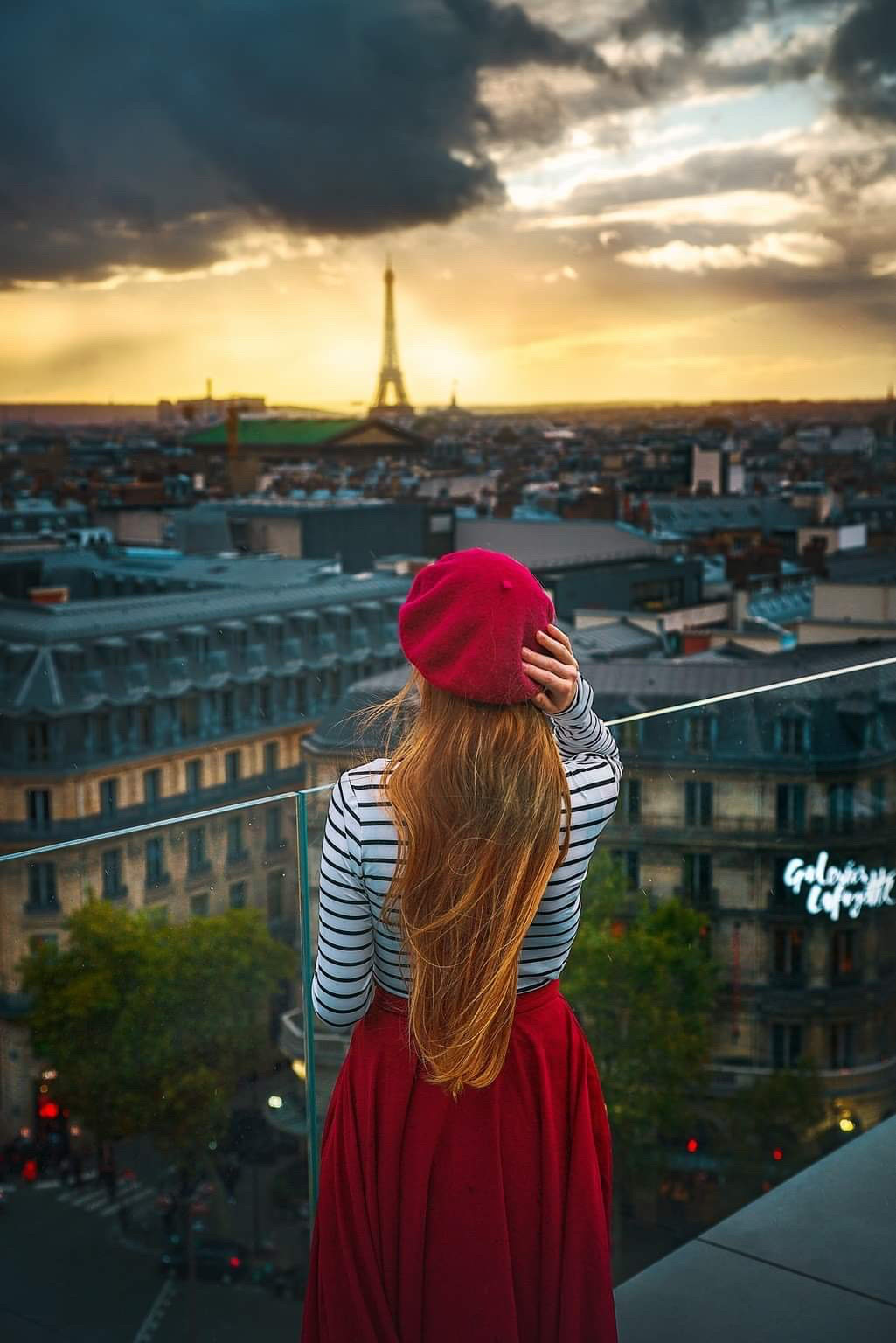 A girl in a red skirt standing at the Free rooftop of Galleries Lafyette overlooking Paris at sunset